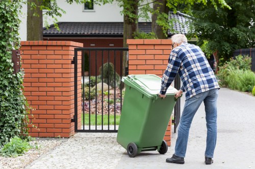 An elderly man dressed in a blue checked shirt and blue jeans is seen leaning forward while pushing a large green wheeled rubbish bin towards a black metal gate on a paved driveway. The bin's lid is closed, and it is positioned close to the gate made of vertical bars and beneath brick pillars, which are part of a garden entrance. Behind the gate, a residential property with a house partially visible, featuring a dark trim roof and white window frames, is surrounded by green hedges and trees. The environment appears tidy and well-maintained, with some shrubbery along the side of the driveway and leafy trees providing shade. This scene illustrates rubbish collection or disposal activity in a suburban area, relevant to professional waste removal services such as those offered by Commercial Waste Slough, serving local businesses and residents in the SL1 postcode area.