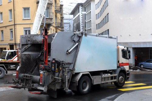A commercial waste collection truck parked on a street in an urban area, with a large, rectangular metal cargo compartment painted in light grey and showing signs of dirt and weathering. The truck is positioned near a pedestrian crossing with yellow markings on the pavement, and the surrounding environment includes multi-storey buildings, some with scaffolding and protective wrapping, indicating ongoing construction or renovation work. On the left, part of a red and white extendable cherry picker or ladder is visible leaning against a building, while on the right, there are parked cars and a driveway leading to a garage or loading bay. The scene is captured during daylight hours, with overcast weather contributing to diffuse lighting. The image reflects the typical urban rubbish collection activity, with possible connections to commercial waste services in Slough, provided by companies like Commercial Waste Slough.