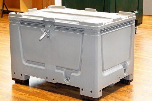 A large, sturdy plastic rubbish skip with a light grey finish, situated indoors on a polished wooden floor. The skip has a rectangular shape with slightly rounded edges and is reinforced with ridges along the sides for added strength. The lid is securely fastened with a metal latch on the front, and the lid appears to be slightly raised at the front edge. The skip’s surface is smooth with a matte texture, and it rests on black plastic feet that elevate it slightly above the floor. In the background, there are green and dark coloured waste containers, indicating a waste management area. The lighting is even, highlighting the skip’s clean, utilitarian design, aligning with professional rubbish removal services offered by Commercial Waste Slough, serving the Slough postcode area. The environment suggests an indoor storage or waste collection point, consistent with commercial waste management facilities. This image visually represents equipment used for collecting and transporting rubbish, relevant to local waste disposal and recycling solutions in the Slough region.
