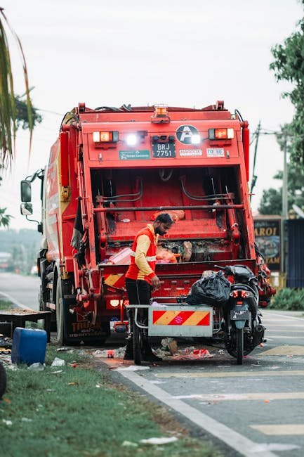 A large, dark green McNeilus rubbish collection truck is positioned on a city street, facing directly towards the viewer. The truck features a sturdy metal front cab with a slightly curved windshield,