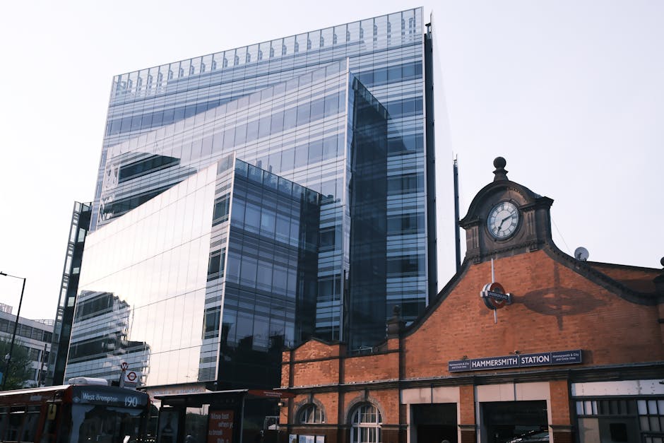 The image shows a modern, multi-storey office building with a glass façade reflecting the surrounding cityscape, contrasting with an adjacent historic brick structure featuring a clock tower. The off